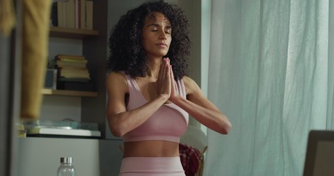 Cinematic shot of young sporty brazilian woman wearing bra and leggings is practicing yoga and meditating seated on mat with online tutorial on laptop at home. 