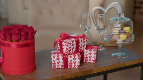Valentine day gift boxes with red hearts wrapping paper on coffee table next to luxury red roses bouquet and gourmet macarons. Valentines romantic surprise with presents and flowers.