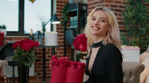 Portrait of smiling valentine day beautiful blonde with heart shape box of roses bouquet in living full of gifts. Happy seductive woman enjoying romantic surprise in room with festive decorations.