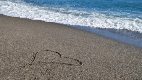 Heart symbol drawing on sand beach with soft blue wave on background. Sea wave with foam wash away a heart drawn on the sand. Valentines day, romantic concept.
