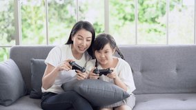 Happy young Asian mother and daughter playing video games online are playing game console and laughing while sitting on sofa in living room at home. Mom and girl celebrating winning.