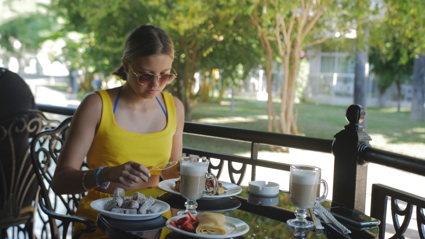 A girl in sunglasses on a summer terrace in a cafe is happy to eat dessert sitting at a cozy table. Coffee and dessert in a cozy summer cafe.