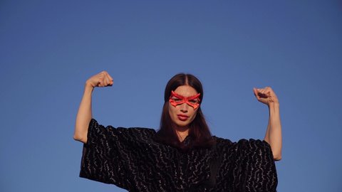 Brunette superhero girl in black dress and red face mask spreading hands with clenched fists and looking at camera on blue sky background. Female power, protest, women rights concept. 4k video footage
