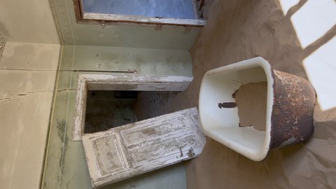 Vertical video. Young woman in robe in abandoned building in bathroom walks bath tub. Ghost town Kolmanskop in desert. Room filled with sand. Old bathtub in ruined house. Girl in white silk underwear