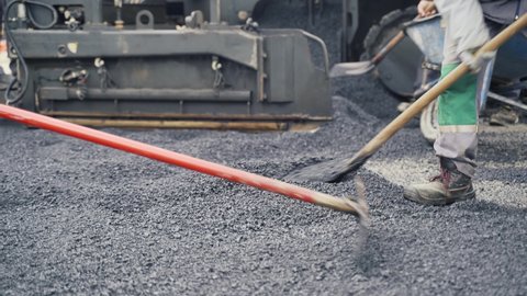 Paving laborers leveling fresh asphalt on a road construction site using asphalt lutes with one of them loading a wheelbarrow in the background.