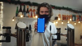 Barman in mask showing blue screen on phone. Bearded man behind bar counter holding smartphone with chroma key. Visiting pubs during coronavirus, green zone restrictions. Bartender in restaurant. - Powered by Shutterstock - Get 15% off with code: PIKWIZARD15
