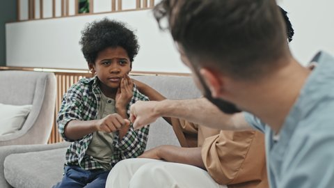 Over-shoulder of cropped male Caucasian doctor wearing scrubs bumping fists with Black ten-year-old boy having toothache, sitting in waiting room at clinic with his female parent, kid smiling