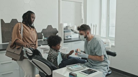 Medium long of male Caucasian doctor showing dentures to ten-year-old boy who sitting in dentist chair and young Black woman standing by his side in modern medical office at daytime