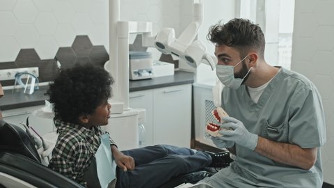 Medium long of male Caucasian doctor entertaining Black ten-year-old boy who sitting in dentist chair with dentures, cropped parent standing by his side in modern medical office at daytime