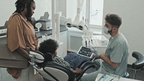 Medium long of male Caucasian doctor showing dental X-ray to ten-year-old boy who sitting in dentist chair and young Black woman standing by his side in modern medical office at daytime