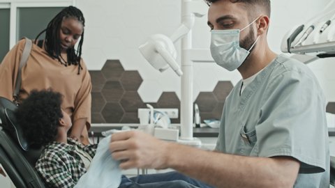 Tilting up and down of male Caucasian doctor wearing scrubs, putting on face mask and gloves before work in his office, Black elementary-aged boy sitting in dentist chair, his mom standing by side