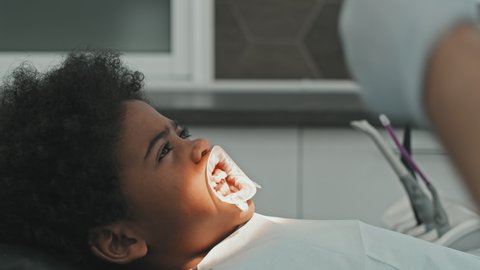 Side-view of scared Black ten-year-old boy lying in dentist chair, wearing dental bib and dental dam, waiting for procedure