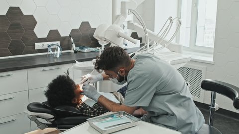 High angle of young male Caucasian doctor wearing mask and gloves, treating cavities of Black boy lying in dentist chair in bright medical office
