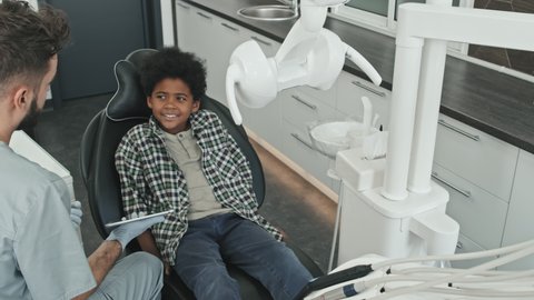 High-angle of Caucasian bearded doctor wearing scrubs and gloves, using tablet computer for oral care education of Black curly-haired little patient sitting in dentist chair at daytime and smiling