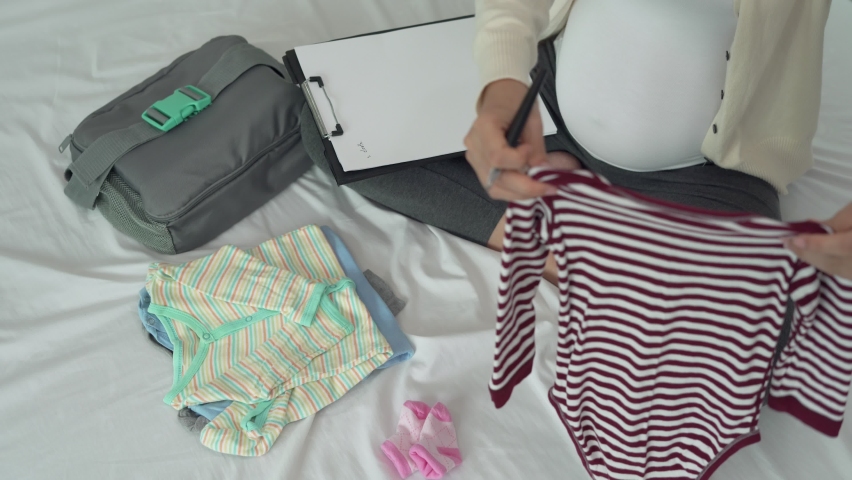 Mother is folding baby clothes to prepare for the upcoming baby. A pregnant woman is checking necessary for newborn. mothers record among of cloth for baby.
