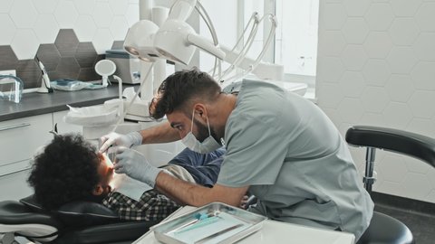 Medium long of young male Caucasian doctor wearing scrubs, mask and gloves, treating cavities of Black boy lying in dentist chair in bright medical office