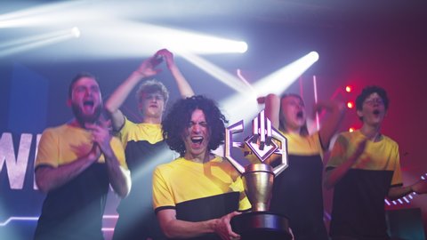 Team of professional gamers esportsman with golden trophy smiling and dancing together while celebrating victory in gaming tournament under bright spotlights