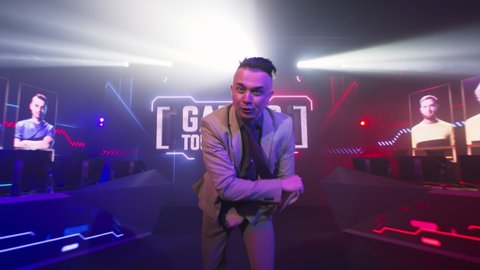 Excited adult announcer speaking to camera at start of professional esports competition on glowing stage with digital screen and neon illumination