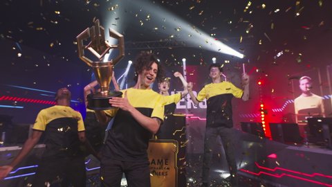 Cheerful diverse esports teammates gamers with golden cup screaming in excitement and celebrating victory in professional gaming championship under falling confetti