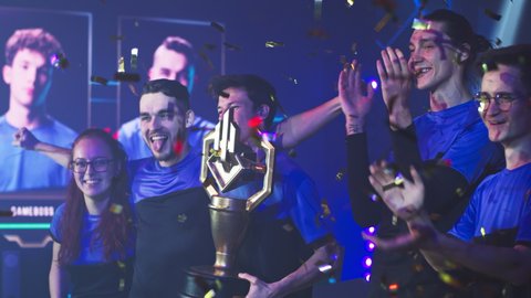 Happy professional esport team of gamers winners with golden cup standing under falling confetti and celebrating victory in professional esports gaming tournament together