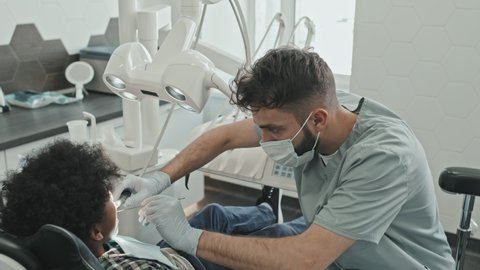 High angle of male Caucasian doctor wearing gloves, mask and scrubs treating Black ten-year-old boy who sitting in dentist chair in modern medical office at daytime