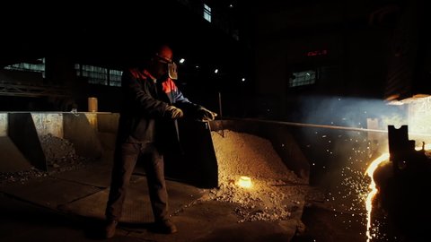 Industrial Worker Manually Creates Molten Metal Stream Using Long Pole Tool. Industrial Worker Handles Molten Metal Production Furnace. Worker On Molten Metal Manufacturing Industrial Factory Line