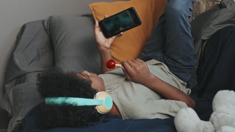 Medium long of ten-year-old Black boy wearing over-ear headphones, lying on bed with legs up on wall in his room at daytime, having lollipop and listening to music on smartphone