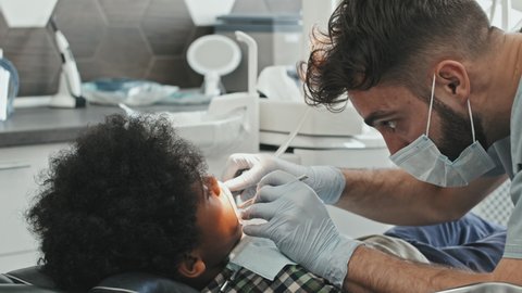 Tracking out of young male Caucasian doctor wearing mask and gloves, taking tool and starting treatment of teeth of Black boy lying in dentist chair in bright clinic