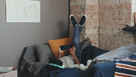 Long shot of ten-year-old Black boy wearing over-ear headphones, lying on bed with legs up on wall in his room at daytime, having lollipop and watching videos on smartphone