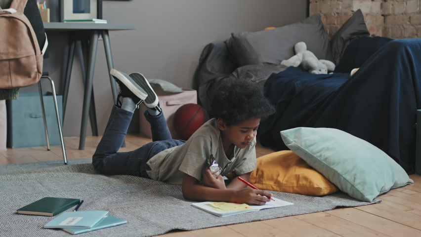 Wide shot of ten-year-old Black boy lying on stomach on floor in his room at daytime, drawing and eating chocolate