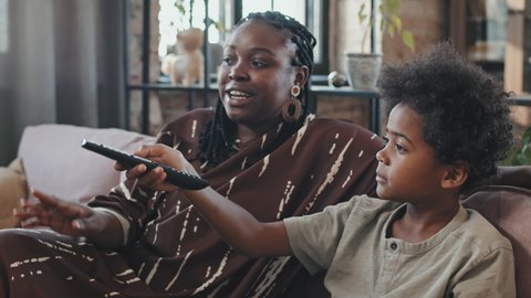 Tilting up and down of young Black woman and her elementary-aged son sitting on couch in living room on weekend, arguing about what to watch on TV and having candies for snack