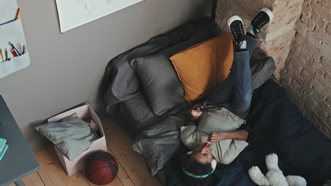 Top view of ten-year-old Black boy wearing over-ear headphones, lying on bed with legs up on wall in his room at daytime, having lollipop and using smartphone