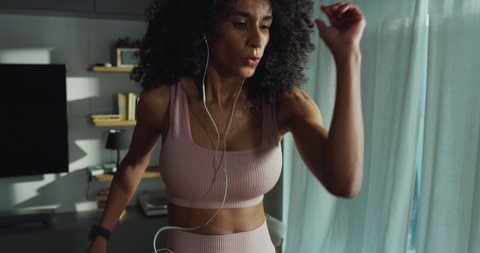 Cinematic shot of young sporty brazilian woman with athletic body wearing bra and leggings is practicing high intensity cardio exercises at home during covid-19 pandemic lockdown.