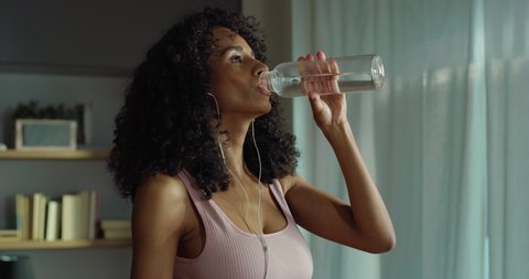 Cinematic shot of young sweaty and thirsty sporty brazilian woman with athletic body wearing bra and leggings drinking fresh cool water from bottle after practicing fitness workout exercises at home.