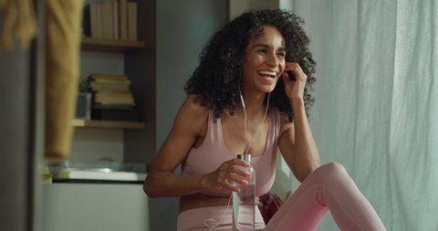 Cinematic shot of happy smiling sporty brazilian woman drinking fresh water from bottle after practicing fitness workout exercises and making technology video call to friend with smartphone at home.