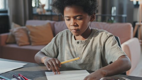 Tilting up of ten-year-old Black boy drawing at desk at home, eating sweets at daytime