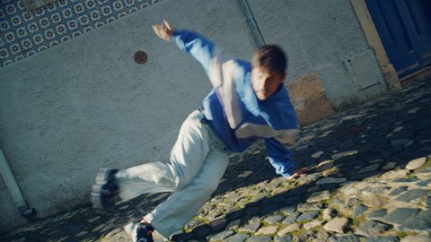 Active and Cheerful Young Adult Latin Man in Stylish Clothes Actively Breakdancing on the Street of an Old Town in a City. Scene Shot in an Urban Environment on an Quiet Small Town Street.
