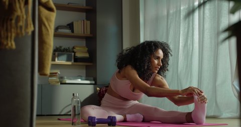 Cinematic shot of young sporty brazilian woman with athletic body wearing bra and leggings is practicing leg stretching exercises on fitness mat at home during covid-19 pandemic lockdown.