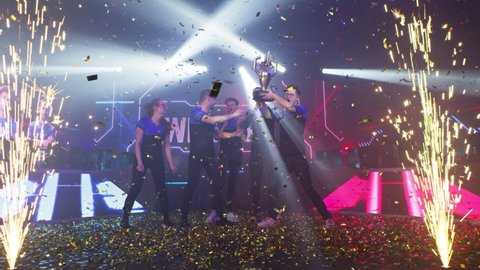 Professional cybersport team of gamers winners with golden trophy hugging and jumping amidst confetti and pyrotechnics while celebrating victory in esports competition