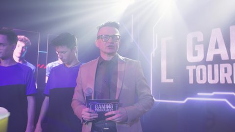 Host man speaking about golden cup while standing near cyber esportsmen at start of gaming tournament