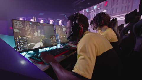 Multiracial young esportsman gamers playing shooter on computers near large monitor with match broadcast during professional gaming competition battle