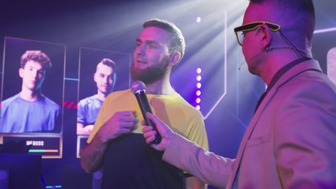 Adult man host with microphone asking questions to bearded gamer esportsman and announcing start of competition during gaming tournament