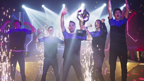 Cybersport team with golden cup jumping and clapping hands near pyrotechnics while celebrating victory in gaming tournament together