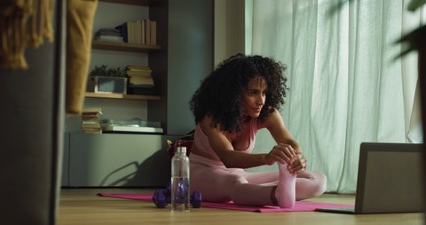 Cinematic shot of young sporty brazilian woman wearing bra and leggings is practicing yoga and meditating seated on mat with online tutorial on laptop at home. 