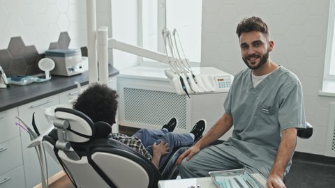 Medium long portrait of bearded Caucasian male doctor wearing scrubs, smiling on camera and posing sitting on dentist stool in his bright office, curly-haired patient sitting in dentist chair