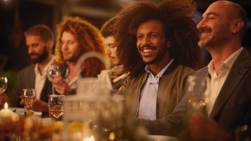 Big Dinner Party with a Small Crowd of Multiethnic Diverse Friends Celebrating at a Restaurant. Beautiful Happy Hosts Propose a Toast and Raise Wine Glasses while Sitting at a Table in the Evening. Royalty-Free Stock Footage #1085993732