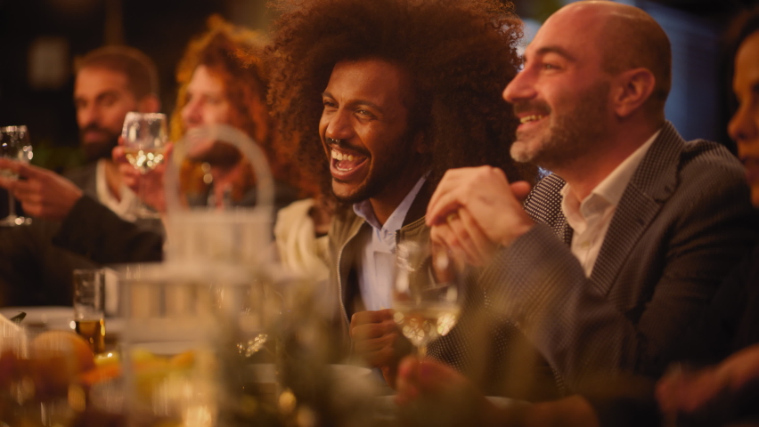 Big Dinner Party with a Small Crowd of Multiethnic Diverse Friends Celebrating at a Restaurant. Beautiful Happy Hosts Propose a Toast and Raise Wine Glasses while Sitting at a Table in the Evening. Royalty-Free Stock Footage #1085993735