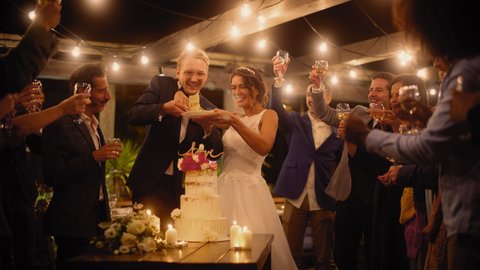 Beautiful Bride and Groom Celebrate Wedding at an Evening Reception Party with Multiethnic Friends. Married Couple Standing at a Dinner Table, Kiss and Cut Wedding Cake.