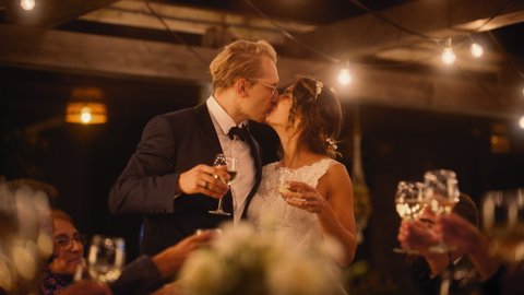 Beautiful Bride and Groom Celebrate Wedding at an Evening Reception Party. Newlyweds Propose a Toast to Happy Marriage, Standing at a Dinner Table with Best Multiethnic Diverse Friends.