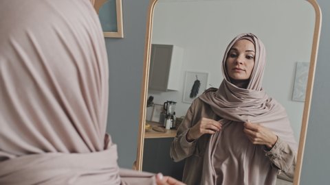 Young beautiful Muslim woman wearing pale pink hijab looking in mirror and smiling while getting dressed at home